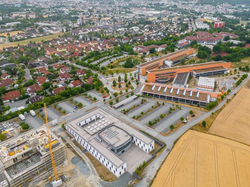 Luftbild eines Geländes mit Neubau und Baukran, dahinter die Stadt