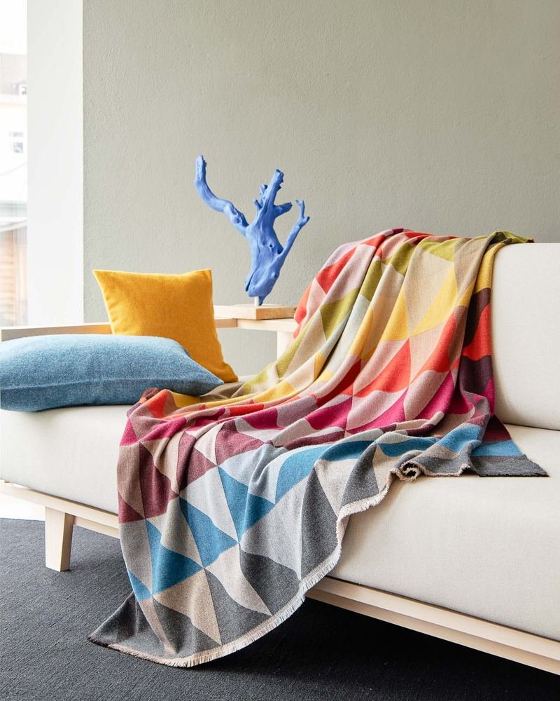 Bunt gemusterte Wohndecke auf hellem Sofa, daneben Kissen und eine blaue Skulptur