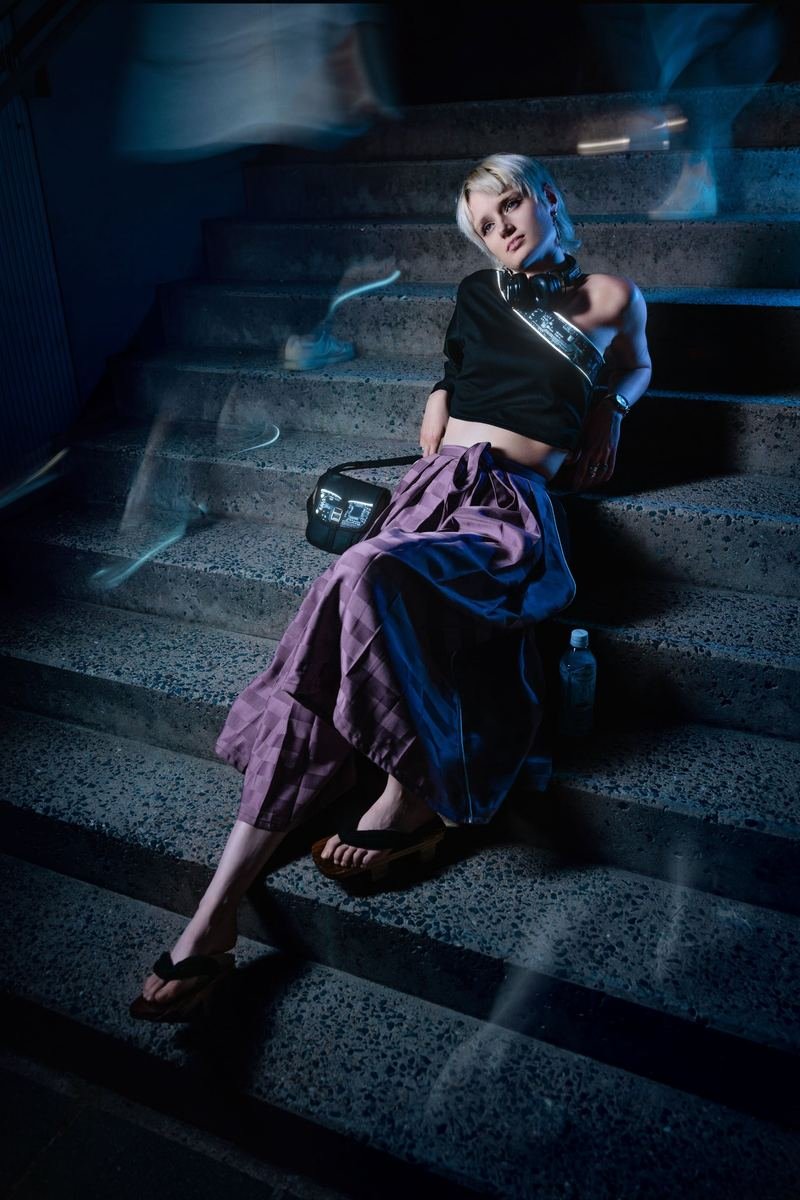 KI-Hybrid-Projekt TSUNAGARI: Model in violettem Rock sitzt nachts auf einer Betontreppe