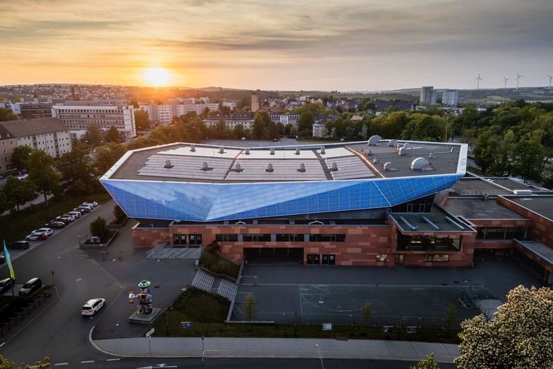 Drohnenaufnahme einer Veranstaltungshalle mit blauem Dach bei Sonnenuntergang über Hof