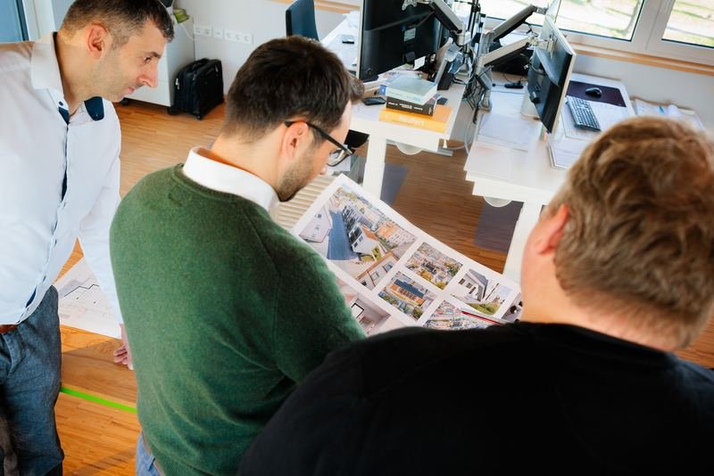 Drei Architekten besprechen am Schreibtisch ausgedruckte Luftbilder und Pläne, von oben fotografiert