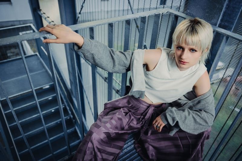 KI-Hybrid-Projekt TSUNAGARI: Model lehnt auf einer Außentreppe, violetter Rock und weißes Oberteil